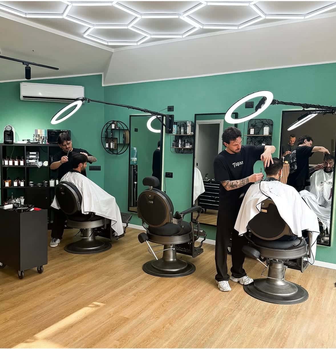 I barbieri al lavoro nella sala verde, coi clienti in poltrona sotto le luci ad anello